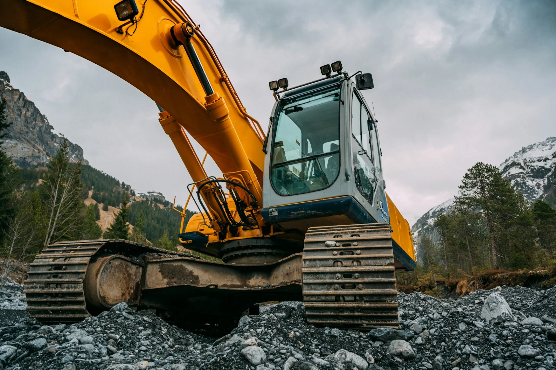 Excavator working on rocky ground beneath evergreen-covered mountains