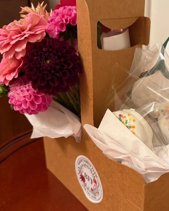 A kraft carrier with a Boght Family Farm sticker, holding pink and burgundy dahlias beside two sprinkled chocolate-covered Oreos