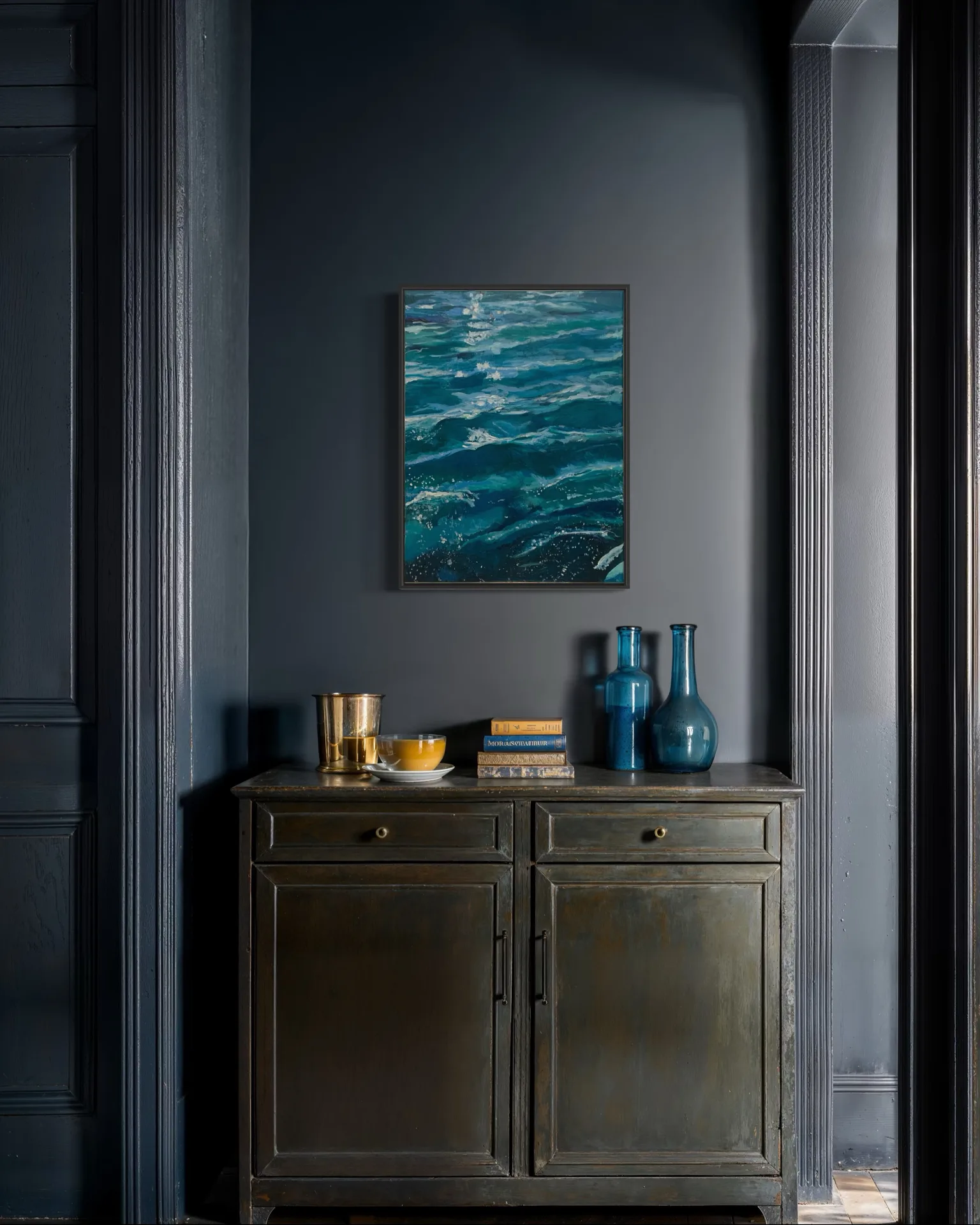 The framed water pastel hung in a dark blue panelled hall above an old painted cabinet with blue glass bottles