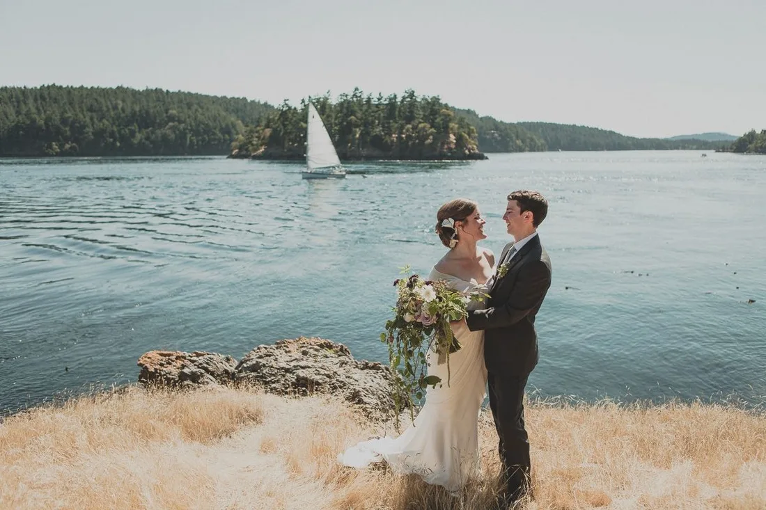 Bride and groom embracing on a rocky shoreline with water and sailboat in the background