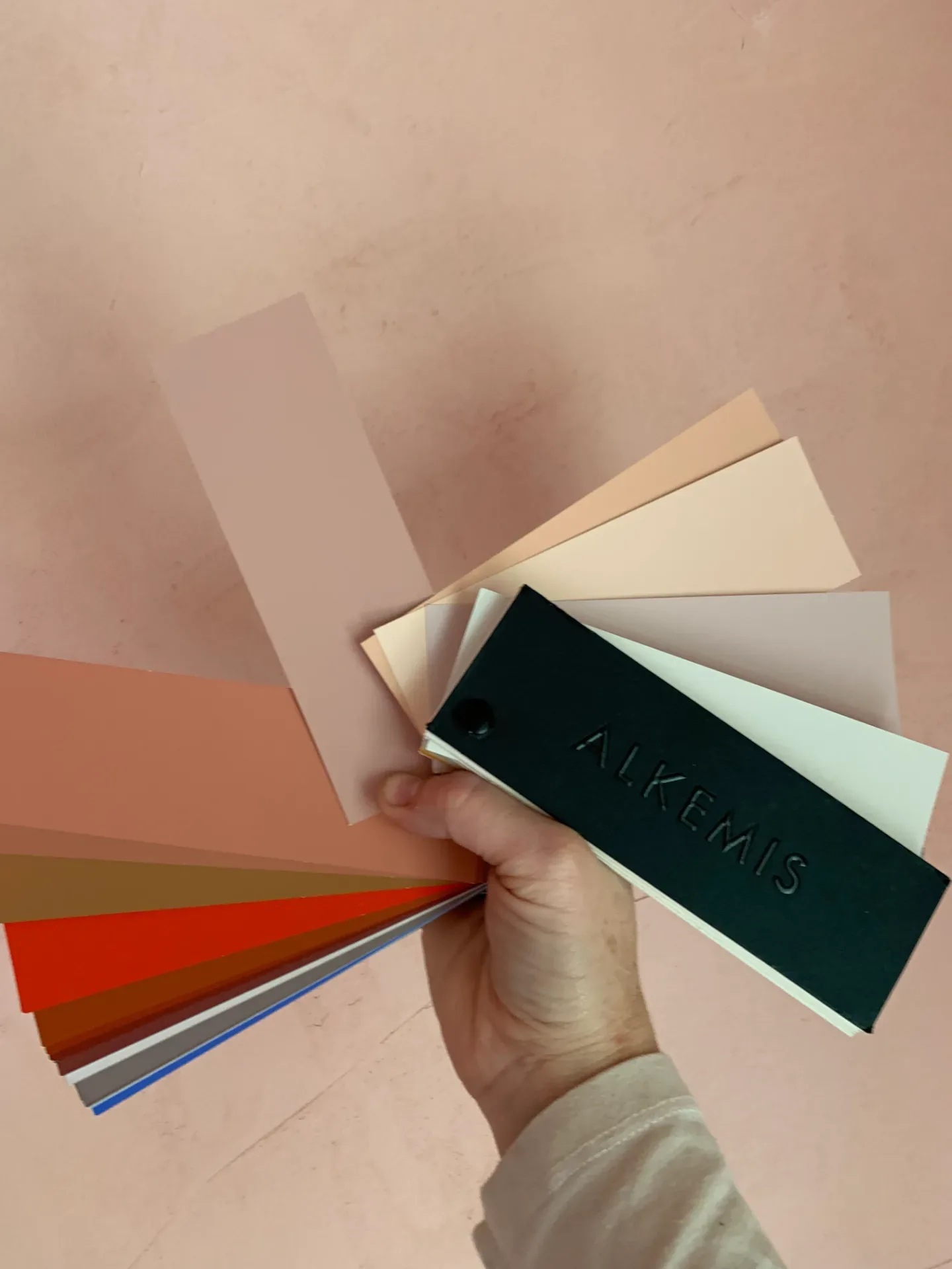A hand holding a fan of paint and material swatches over drawings on a wooden desk