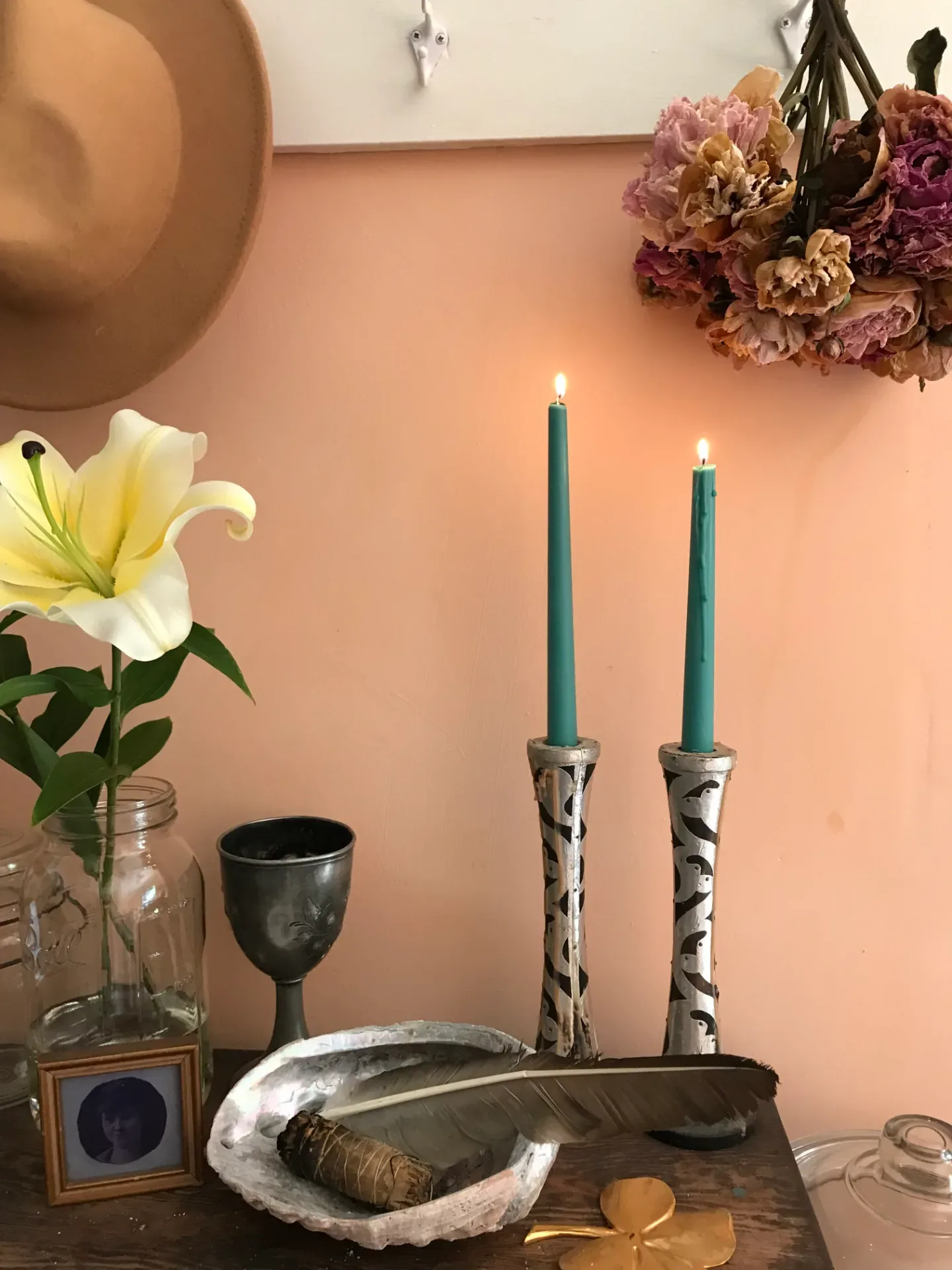Two teal taper candles burning in hand-painted silver candlesticks, beside dried peonies, a white lily, and a pewter goblet against a peach-colored wall