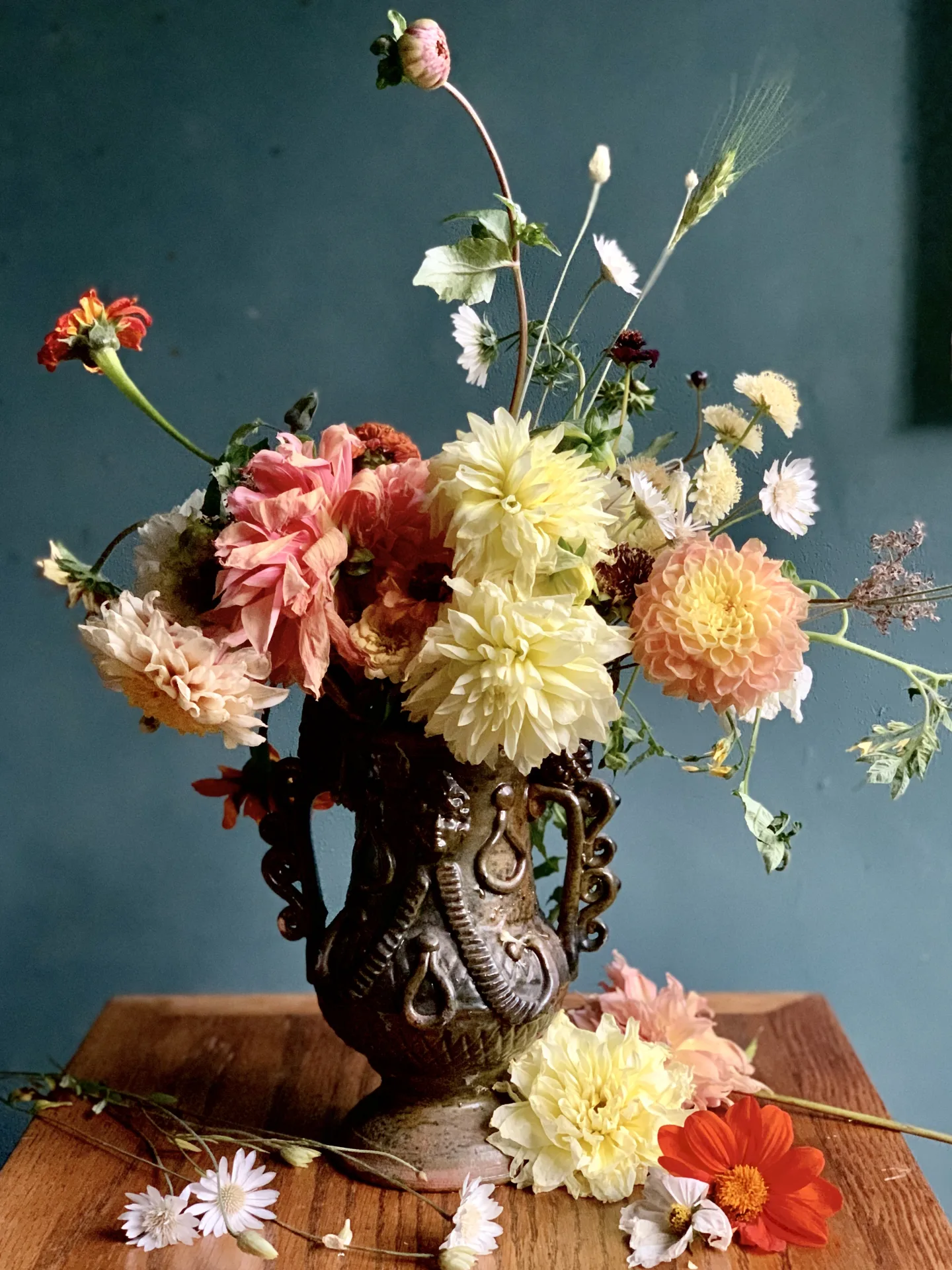 A Dutch still-life style arrangement of cream and peach dahlias, zinnias and daisies in a dark handmade pottery urn on a wood table