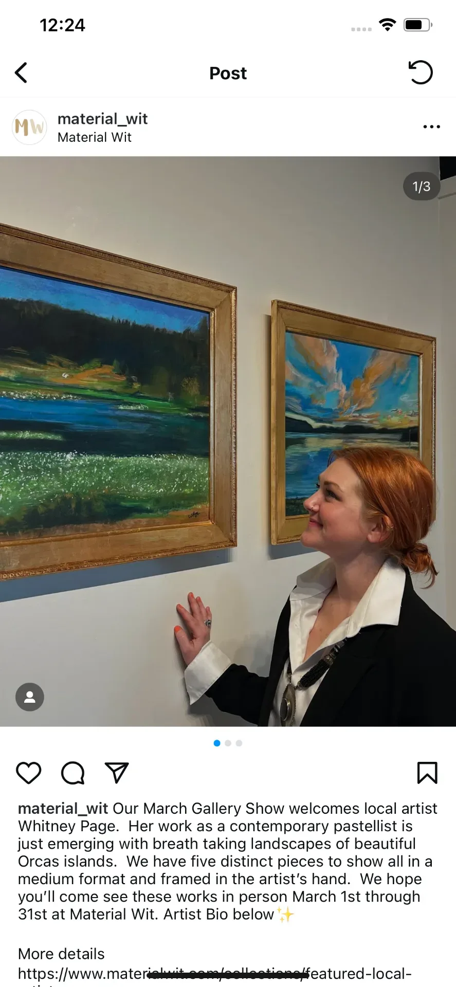 Whitney Page, red hair pinned back and wearing a black jacket, smiling up at two of her framed pastel landscapes hung in gold frames on a gallery wall