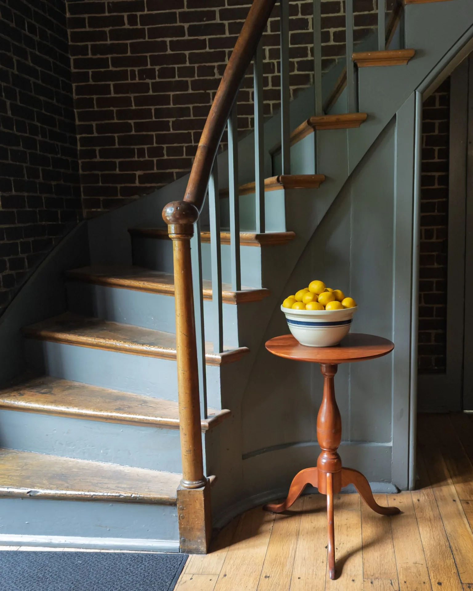 An old staircase with worn fir treads and a turned newel post against exposed brick, a bowl of lemons on a cherry candlestand