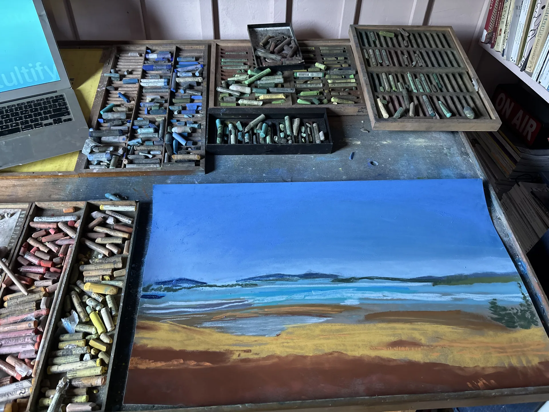 Trays of soft pastel sticks on a studio worktable beside an unfinished pastel of a shoreline on blue paper