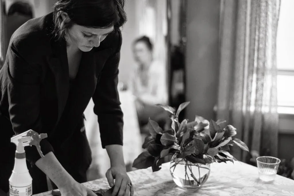 Whitney Page leaning over a table of cut foliage and a glass bowl during a floral class, a student watching from behind