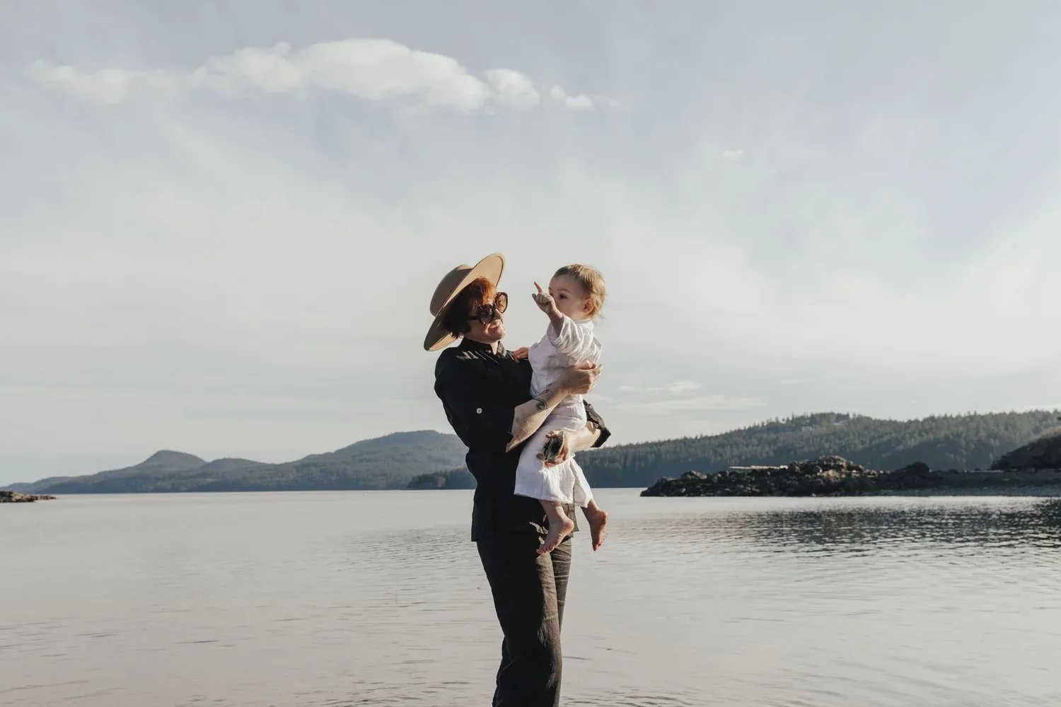Whitney Page holding her child on a calm shoreline with forested islands in the distance
