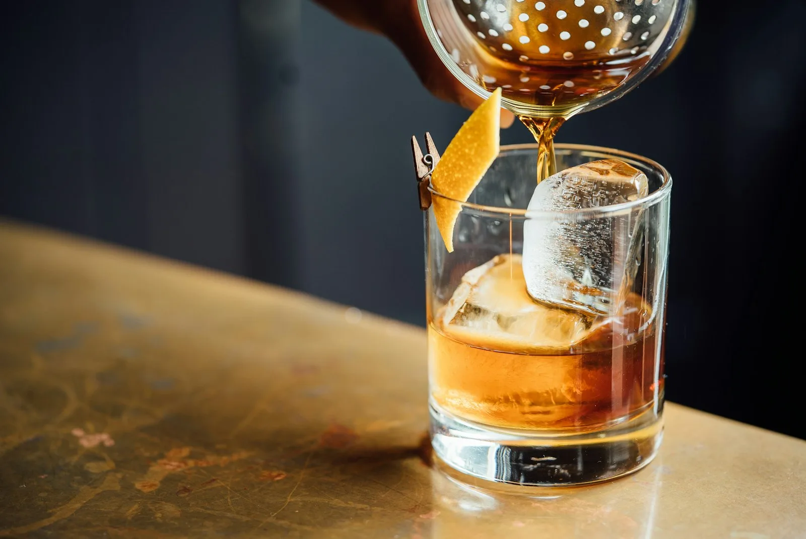 A cocktail being strained over a large ice cube with orange peel