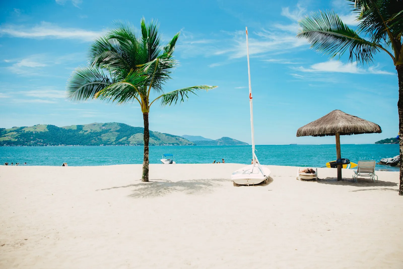 A palm tree, sailboat and thatched umbrella on a bright white beach