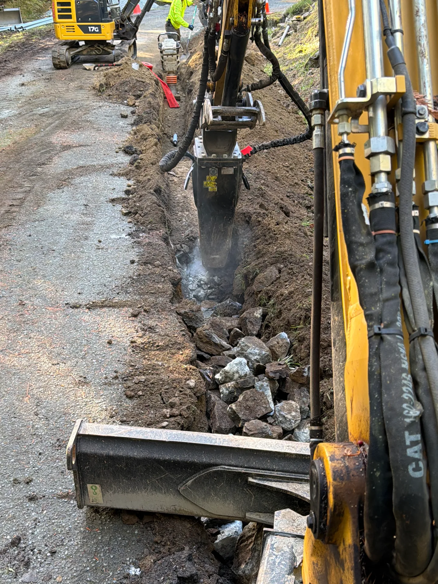 Excavator with a hydraulic breaker hammering rock in a utility trench beside a gravel road