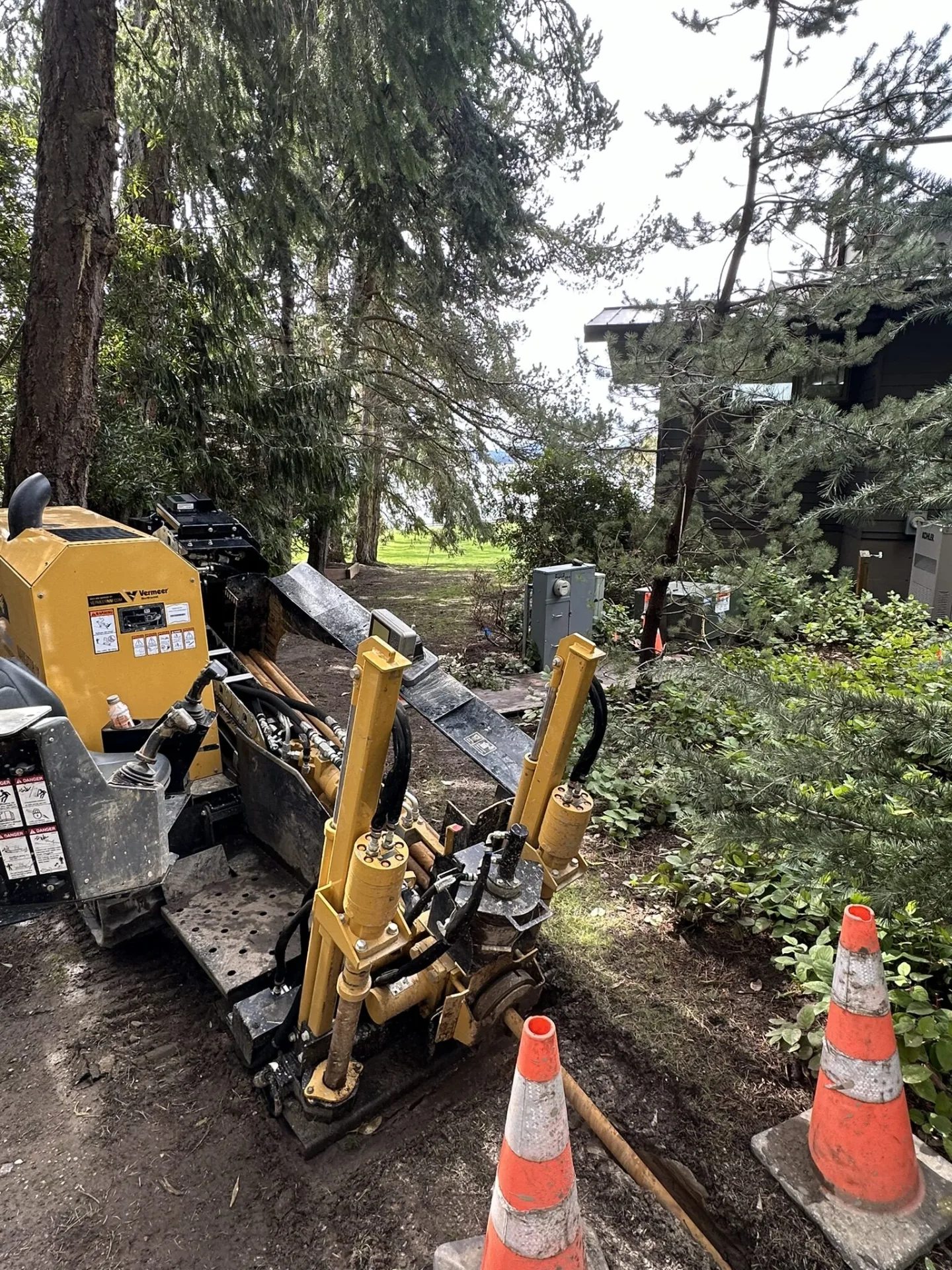 Directional drill rig boring beside a house and its utility meters, among tall evergreens