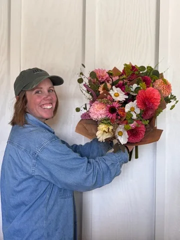 A smiling woman in a green cap holding a bouquet of pink and orange dahlias, daisies and foliage against a white wall