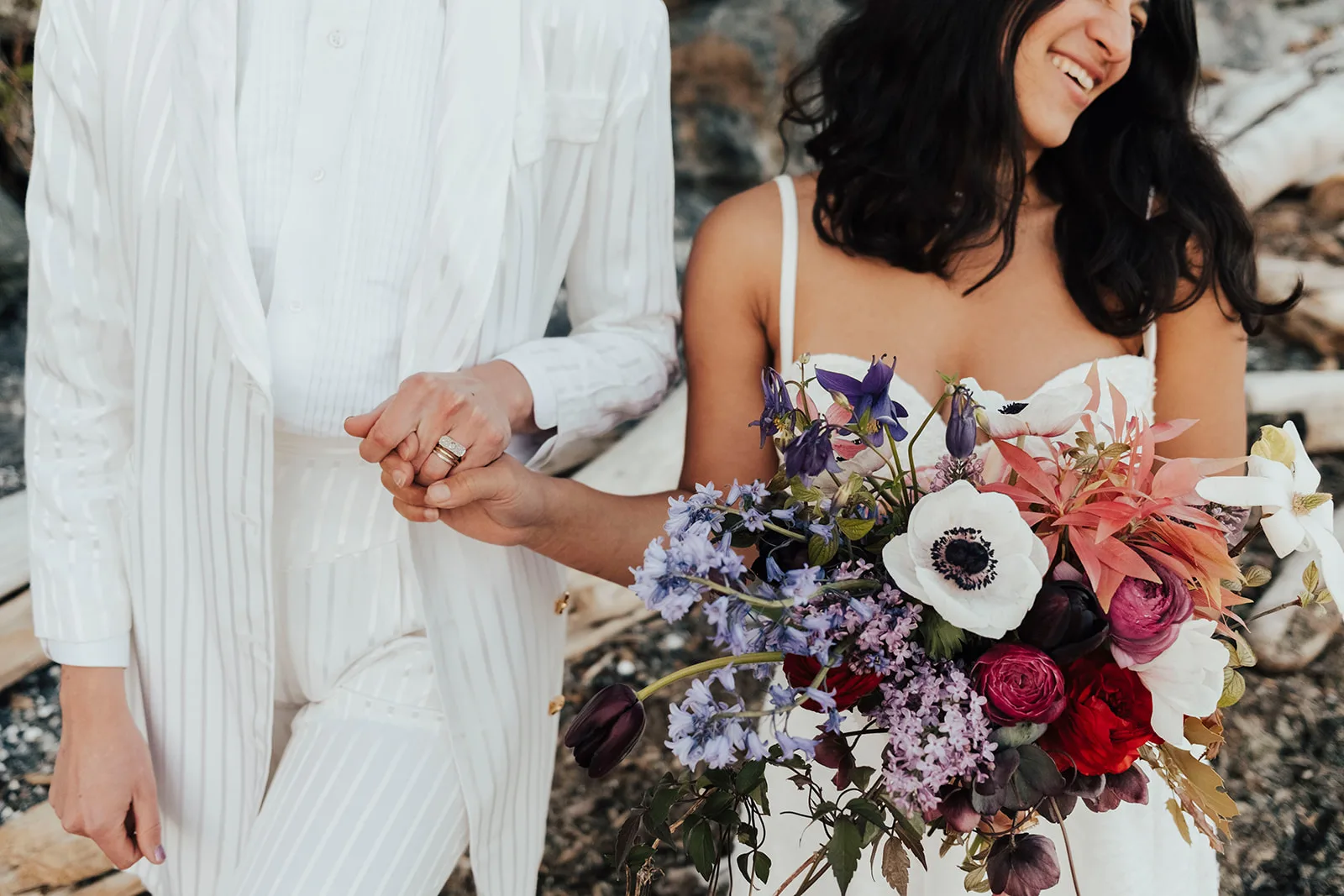 Two brides holding hands on the beach, one carrying a lush bouquet of white anemones, lilac, bluebells, columbine, ranunculus and coral foliage