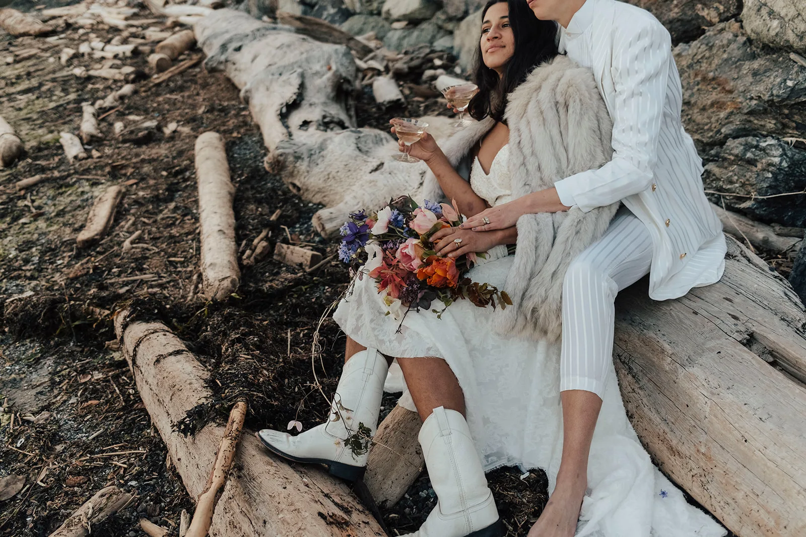 Couple sitting on driftwood with champagne coupes, a bouquet of tulips, roses and delphinium resting in the bride’s lap