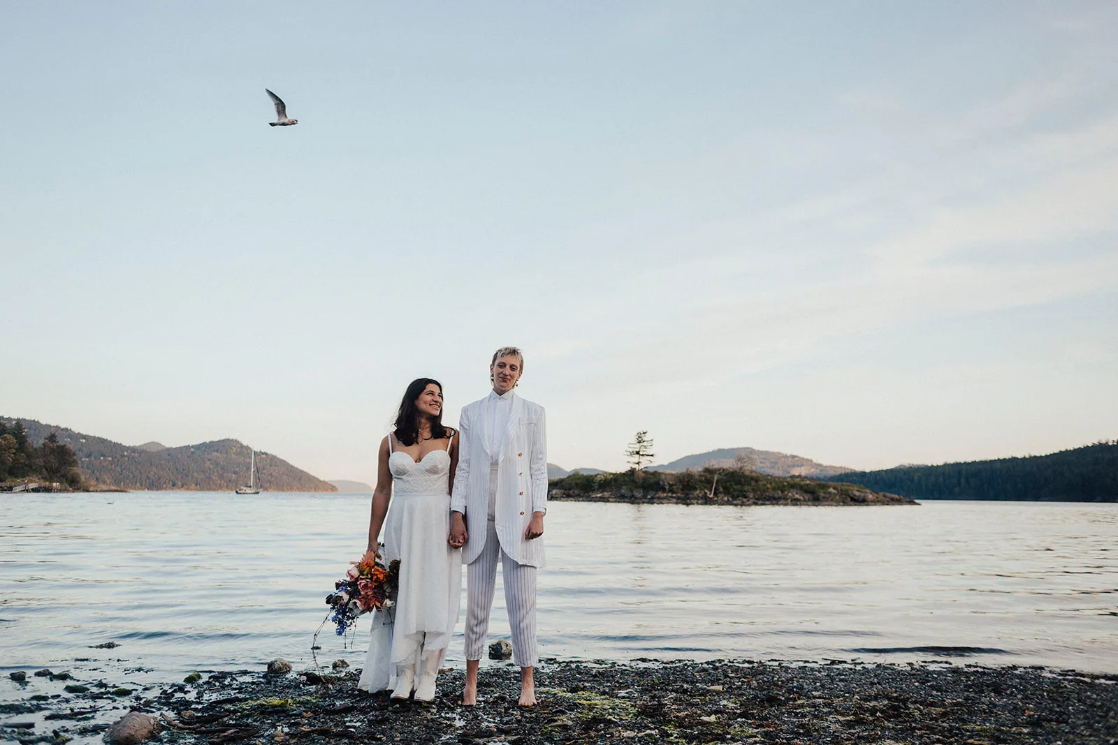 Couple holding hands at the water’s edge on Orcas Island, a trailing bouquet in hand and islands and a sailboat behind them