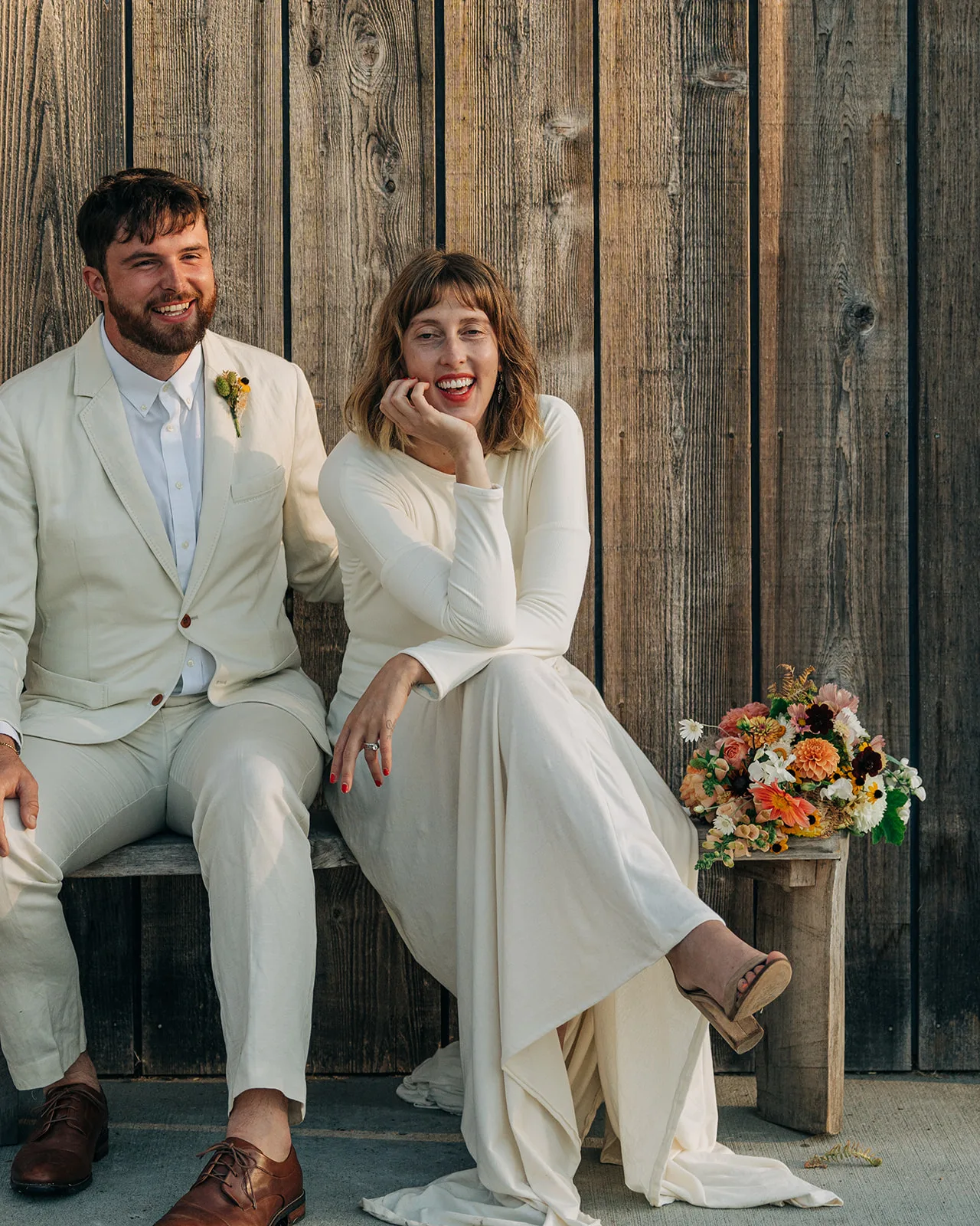 Laughing couple in cream outfits seated on a wooden bench, her bouquet of dahlias and wildflowers beside them